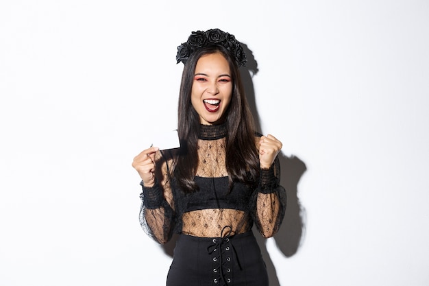 Cheerful asian woman in gothic lace dress and black wreath showing credit card and rejoicing, standing over white background.