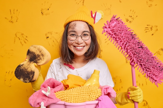 La donna asiatica allegra tiene le attrezzature per la pulizia essendo di buon umore dopo aver riordinato la stanza indossa il cappello occhiali rotondi posa vicino al cesto della biancheria imbrattato di sporco isolato sul muro giallo