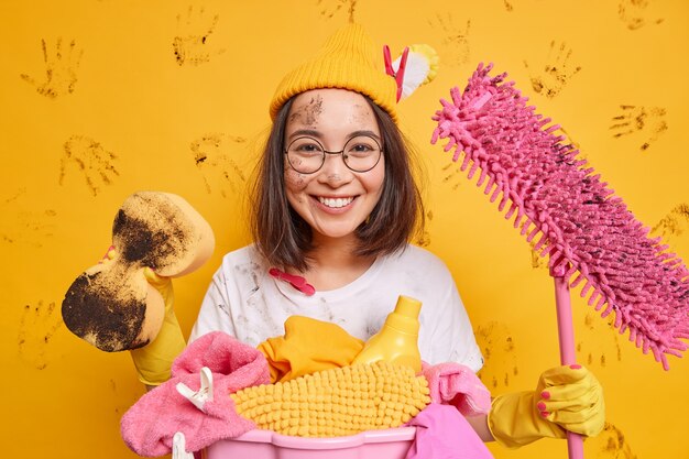 Cheerful Asian woman holds cleaning equipment being in good mood after tidying up room wears hat round spectacles poses near laundry basket smeared with dirt isolated over yellow wall