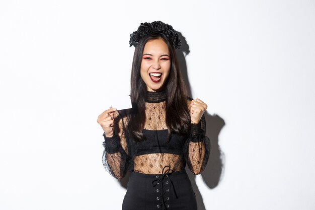 ゴシックレースのドレスと黒の花輪の陽気なアジアの女性は クレジットカードを表示し 白い背景の上に立って喜んでいます 無料の写真