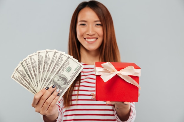 Жизнерадостная азиатская женщина в свитере представляя деньги и подарок на камере над серой предпосылкой