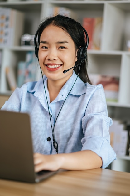 Веселая азиатская женщина носит гарнитуру улыбается и использует потоковую конференцию для видеозвонков на ноутбуке для работы в Интернете во время карантина, самоизоляция covid-19 дома, концепция работы из дома