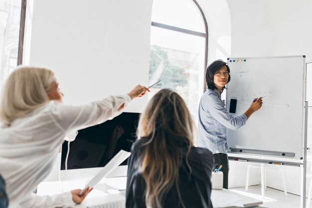 Cheerful asian young man in glasses drawing diagram on flipchart