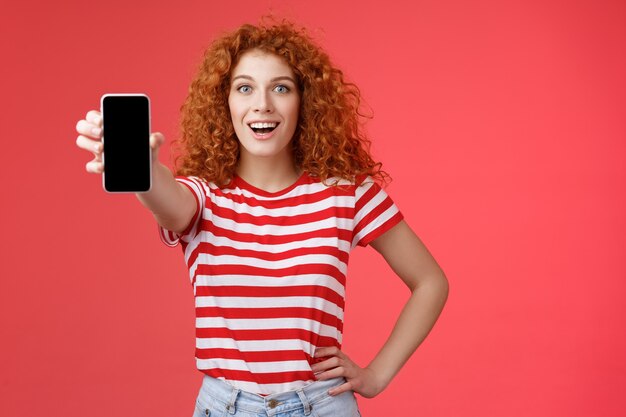 Веселая привлекательная харизматичная европейская рыжая девушка с кудрявой прической показывает экран смартфона, счастливо улыбаясь, продвигая советы по приложениям, рекомендую хорошее приложение на странице в социальных сетях на красном фоне.