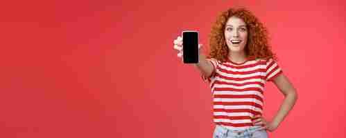 Free photo cheerful attractive charismatic european redhead girl curly hairstyle show smartphone screen smiling