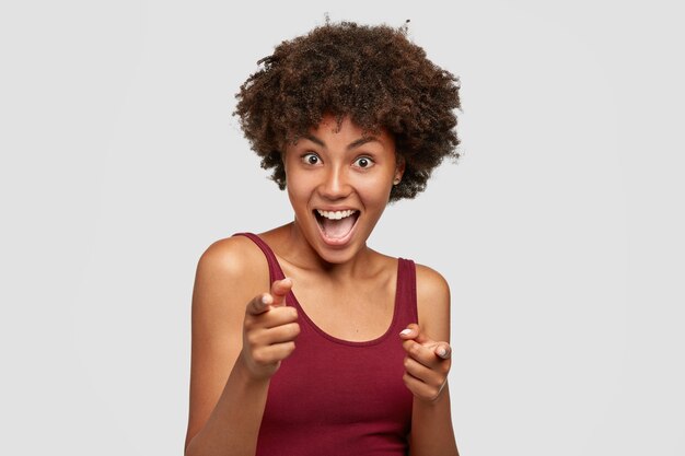 Cheerful attractive dark skinned young girlfriend points to you, has joyful look at camera