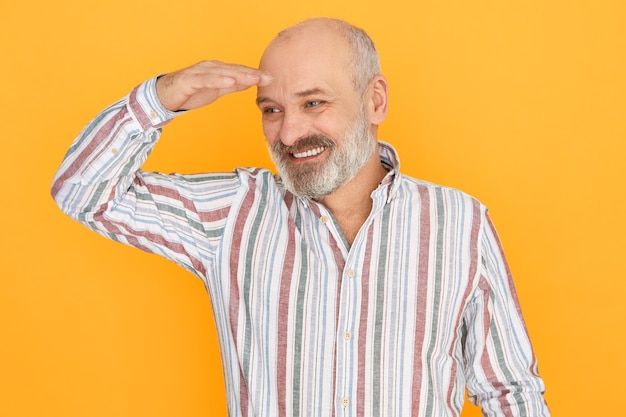 Cheerful attractive elderly male with gray beard and bald head posing isolated keeping hand over his eyes to protect himself
