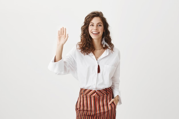 Веселая привлекательная женщина машет поднятой рукой, чтобы поздороваться, дружеское приветствие