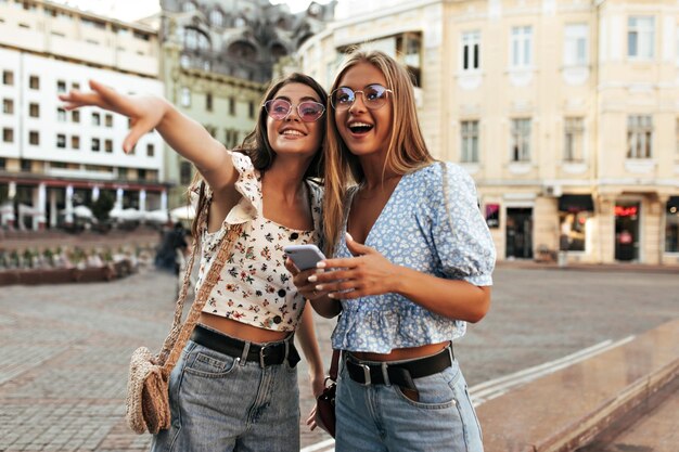 세련된 여름 의상을 입은 쾌활한 매력적인 여성들은 화려한 선글라스를 쓴 흥분한 금발 소녀와 갈색 머리 소녀들이 야외에서 포즈를 취하고 미소를 짓고 놀라움으로 거리를 바라봅니다.