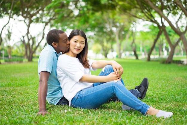 Cheerful attractive young Chinese girl kissed by boyfriend sitting on grass