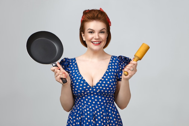 Free photo cheerful attractive young lady wearing retro hairstyle and low cut dress holding wooden pestle in one hand and frying pan in other, bragging of her new cooking utensils, smiling happily at camera