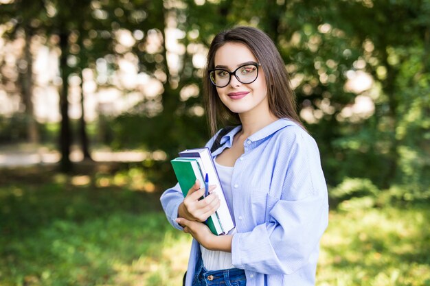 Cheerful attractive young woman with books standing and smiling in park