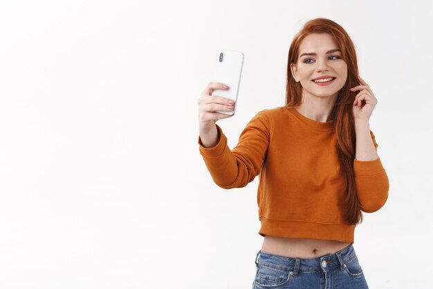 Cheerful attractive young woman with red hair taking selfie before going party, make post internet, hold smartphone looking mobile at front