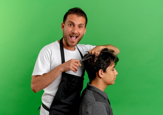Cheerful barber man in apron cutting hair with scissors of satisfied client standing over green wall