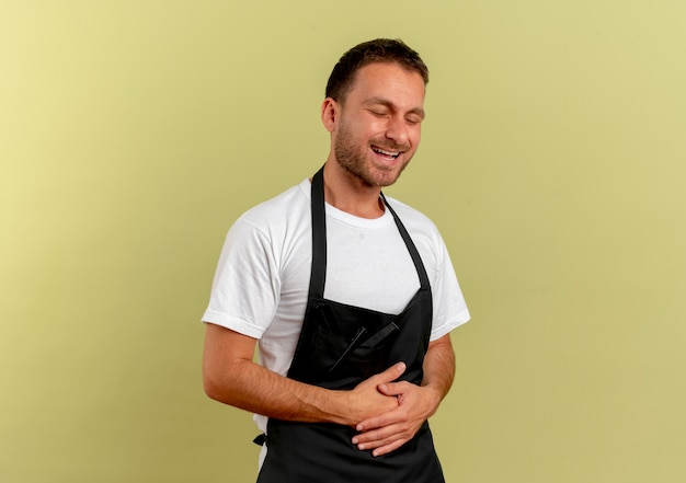 Cheerful barber man in apron laughing out touching his belly standing over light wall