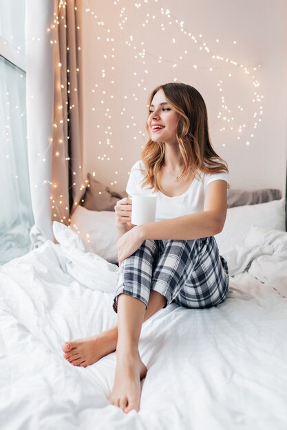 Cheerful barefooted girl sitting on bed and looking at window. Indoor portrait of pleasant caucasian lady in pajama drinking tea.