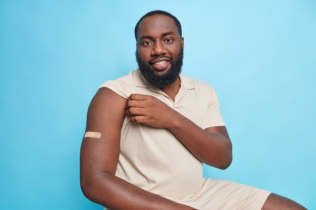 Cheerful bearded adult man gets vaccinated according to schedule in clinic shows arm with adhesive plaster sits against blue wall