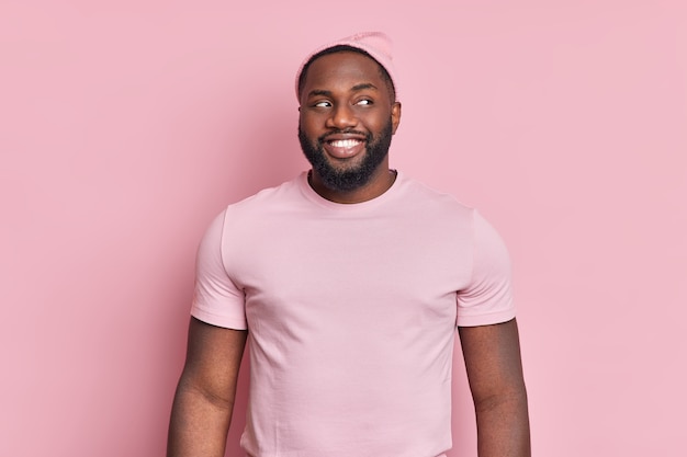 Cheerful bearded black man smiles broady looks curiously aside has white even teeth wears hat and t shirt in one tone with wall