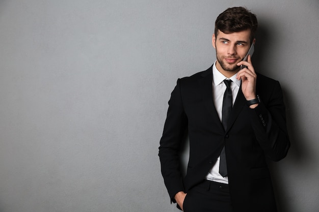 Cheerful bearded guy in black suit speaking on mobile phone, looking aside