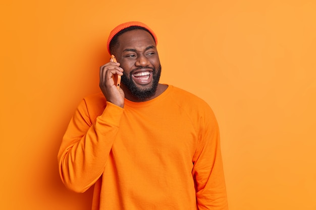 Cheerful bearded guy makes phone call smiles broadly has white teeth dressed in orange sweater and hat looks aside happily discusses plans for weekends