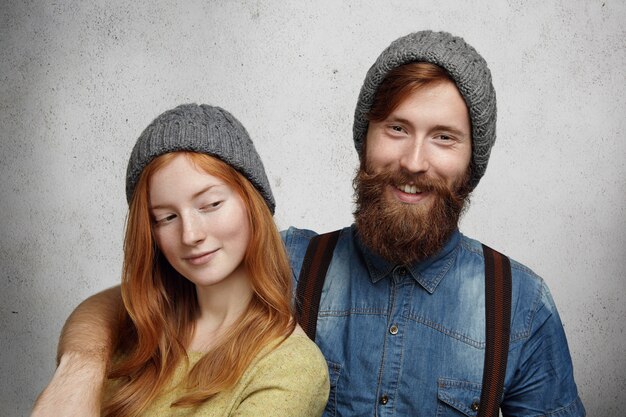 Cheerful bearded hipster in denim shirt laughing while hugging his beautiful redhead girlfriend who is looking at him with sly mysterious smile.