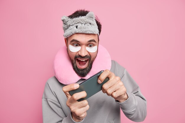 陽気なあごひげを生やした男は、寝る前にスマートフォンでビデオゲームをプレイします。スリープマスクを着用し、現代のテクノロジーに夢中になっている首の周りの快適な枕を着用します。