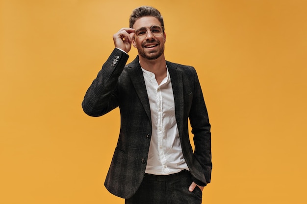 Cheerful bearded man in stylish checkered jacket and white shirt puts on round eyeglasses and poses on orange background