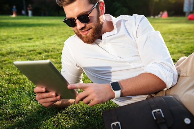 Cheerful bearded man in sunglasses lying on grass outdoors and using his tablet computer