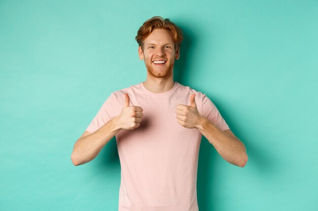 Cheerful bearded man with red hair showing thumbs-up, like and approve something, praising promo, standing against turquoise background.