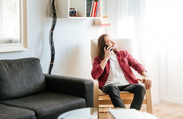 Cheerful bearded young man sitting and talking on cell phone in armchair at home