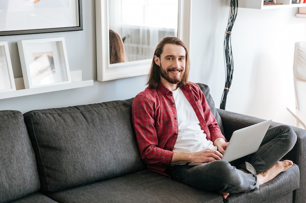 Веселый бородатый молодой человек, используя ноутбук на диване у себя дома
