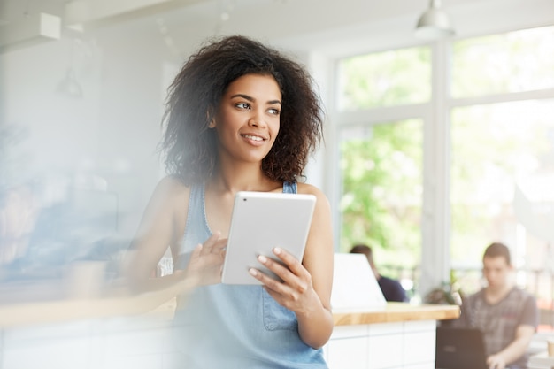 Cheerful beautiful african female student resting in cafe smiling looking in side holding tablet.