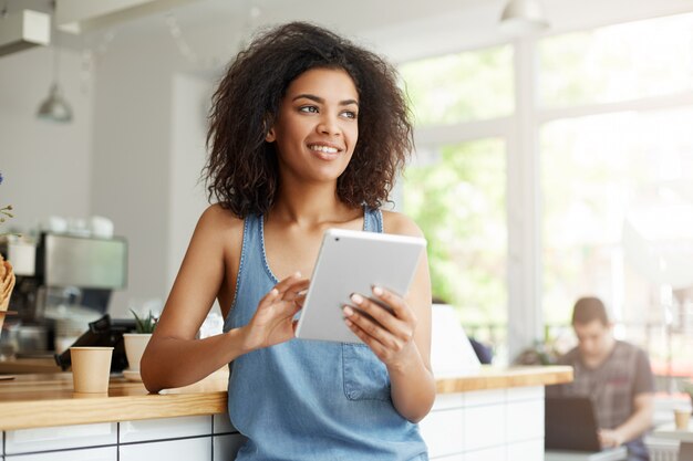 Cheerful beautiful african female student resting in cafe smiling looking in side holding tablet.