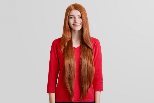 Foto gratuita allegra bella donna con i capelli rossi molto lunghi, la faccia lentigginosa, vestita in maglione rosso casual, posa contro il muro bianco, ha un piacevole sorriso gentile. concetto di positività, bellezza e stile