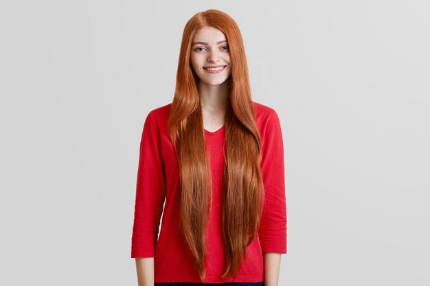 Cheerful beautiful female with very long red hair, freckled face, dressed in casual red sweater, poses against white wall, has pleasant gentle smile. Positivity, beauty and style concept