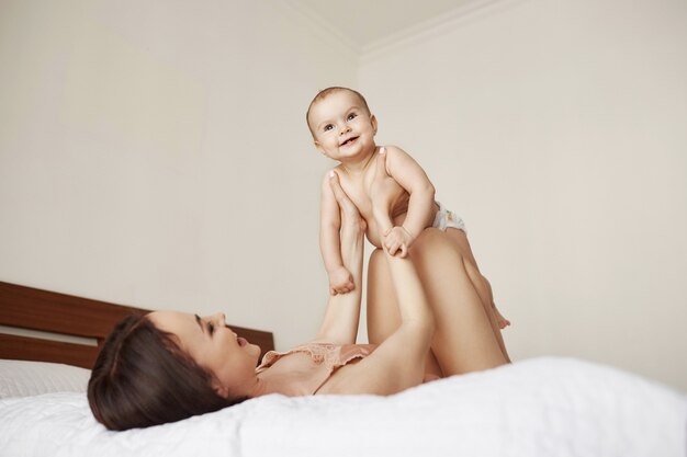 밝고 아름다운 엄마와 그녀의 아기가 집에서 침대에 누워 기쁨을 웃고 연주.