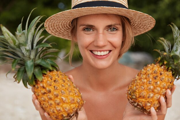 Cheerful beautiful smiling woman with attractive look, broad smile, wears summer straw hat, holds two pineapples, going to make juice, enjoys good rest in tropical country. Female tourist with fruits