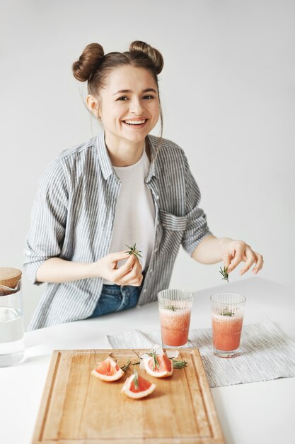 Жизнерадостная красивая женщина усмехаясь украшающ smoothie вытрезвителя грейпфрута с розмариновым маслом над белой стеной. Здоровое питание.