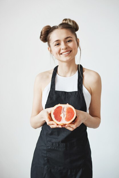 Cheerful beautiful woman with buns in apron smiling holding half of grapefruit in hands over white wall.