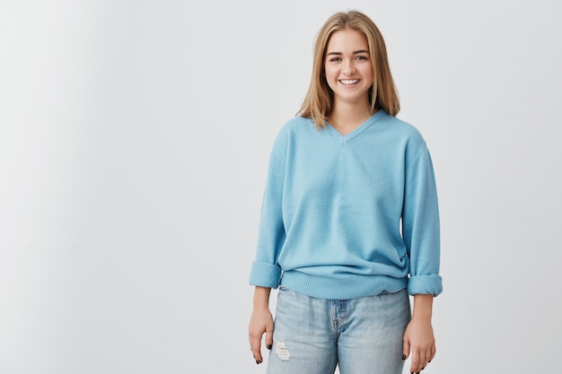 Cheerful beautiful woman with straight fair hair having dark charming eyes and engaging smile posing at studio . Pretty smiling girl wearing blue sweater and jeans.