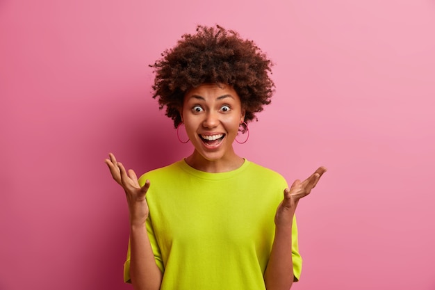 Cheerful beautiful young Afro American woman raises hands reacts positively on great news dressed in casual clothes isolated over pink wall