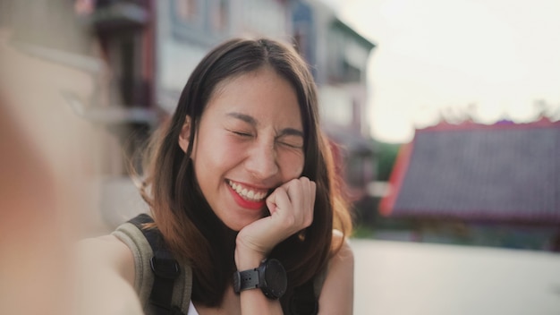 Cheerful beautiful young Asian backpacker blogger woman using smartphone taking selfie 