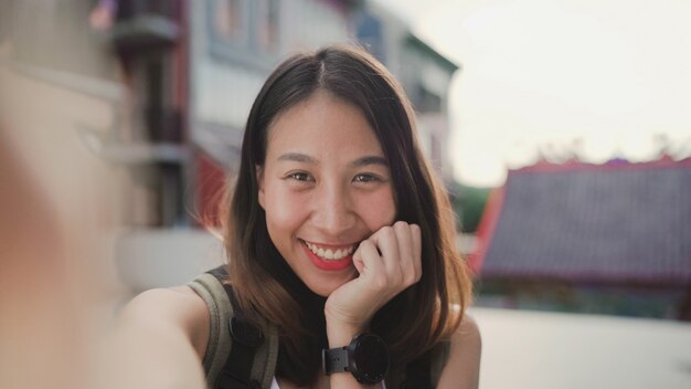 Cheerful beautiful young Asian backpacker blogger woman using smartphone taking selfie 