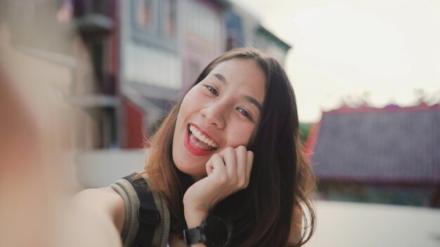 Cheerful beautiful young Asian backpacker blogger woman using smartphone taking selfie