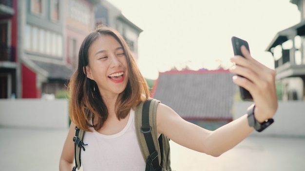 Cheerful beautiful young Asian backpacker blogger woman using smartphone taking selfie 