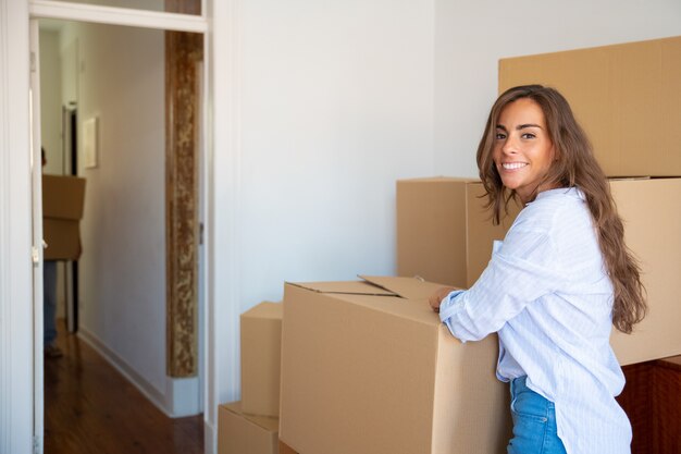 Cheerful beautiful young Hispanic woman unpacking stuff in her new apartment, standing near stacks of carton boxes, 