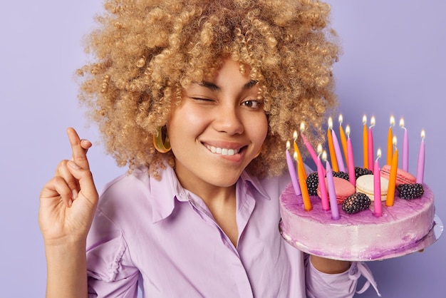 Free photo cheerful birthday girl crosses fingers makes wish before blowing out candles on festive cake winks eye and bites lips hopes dreams come true isolated over purple background holiday concept