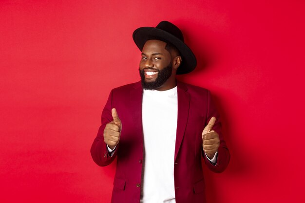 Cheerful Black man having fun on party, showing thumbs up in approval, smiling and liking something, standing against red background