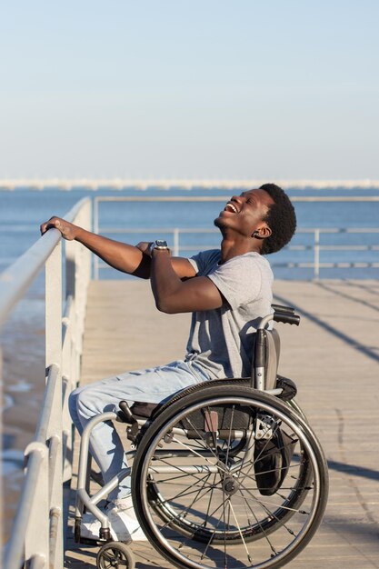 Веселый темнокожий мужчина сидит в инвалидной коляске на берегу моря в солнечный день. Вид сбоку на уверенного в себе парня с инвалидностью, который держит перила рукой и счастливо смеется. Инвалидность, концепция отношения.