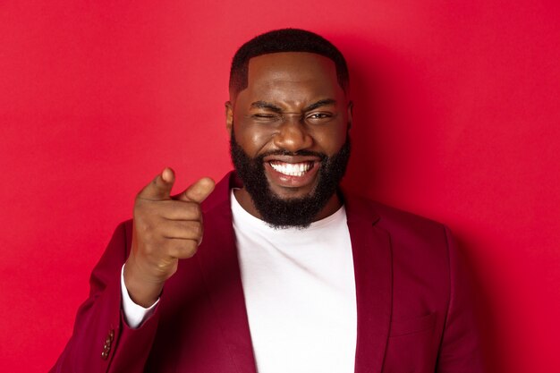 Cheerful Black man with beard wearing party clothes, pointing finger at camera and winking